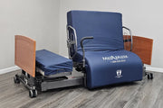 A MedMizer adjustable hospital bed with a navy blue padded mattress and backrest, featuring a wooden headboard and footboard, set on a gray wood-look floor against a light gray wall.