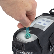 A close-up of a hand inserting a translucent teal tube into a white device attached to a black bag, shown against a plain white background.