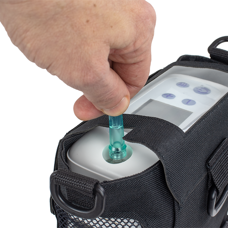 A close-up of a hand inserting a translucent teal tube into a white device attached to a black bag, shown against a plain white background.