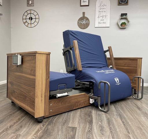 An electric hospital-style bed with a dark blue padded mattress and frame, set in a bedroom or therapy room with wood flooring and wall decor.