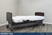 A medical bed frame in Obsidian Oak finish with a white mattress and fitted sheet, shown in a room corner with wood-look flooring. A blue banner at the bottom reads 'Obsidian Oak' and 'MED MIZER'.