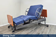 Hospital bed with blue cover and dining chair attachment on a wooden floor.