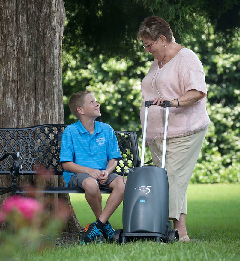 Woman pushing a concentrator next to a boy sitting on a bench in a park.