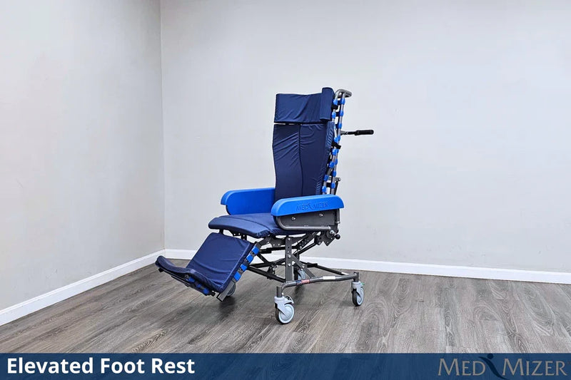 A blue and black medical wheelchair with an elevated foot rest, shown in a clean indoor room with light gray walls and wood-look flooring. A dark blue banner at the bottom reads 'Elevated Foot Rest' and 'MED MIZER'.