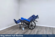 Blue reclining wheelchair with mag wheels in a room with gray walls and wooden floor.