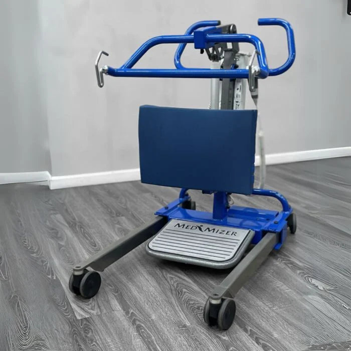 A blue and gray motorized sit to stand with a padded seat, armrests, and a branded footrest labeled 'MedMIZER', shown on a gray wood-look floor against a light gray wall.