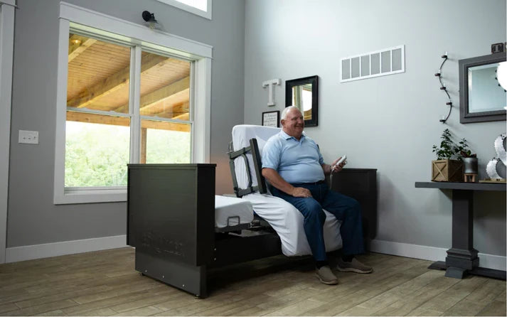 A man sits on a black adjustable bed frame in a modern bedroom with gray walls and wood floors, holding a smartphone.