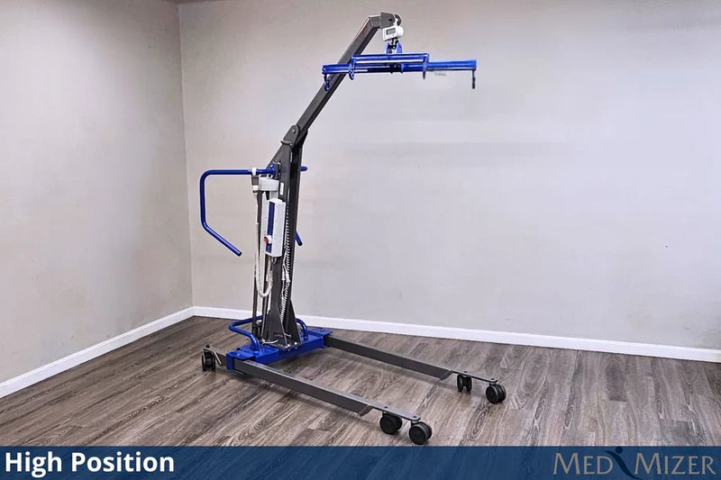 A high-position medical lift chair with a blue base and arm, gray frame, and black wheels, shown in a room corner with gray walls and wood-look flooring. A blue banner at the bottom reads 'High Position' and 'MED MIZER'.