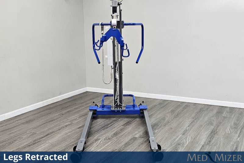A blue and silver medical lift with a wheeled base stands in a room corner with gray wood-look flooring and light gray walls. The lift is shown with its legs retracted, and text overlays at the bottom read 'Legs Retracted' and 'MED MIZER'.