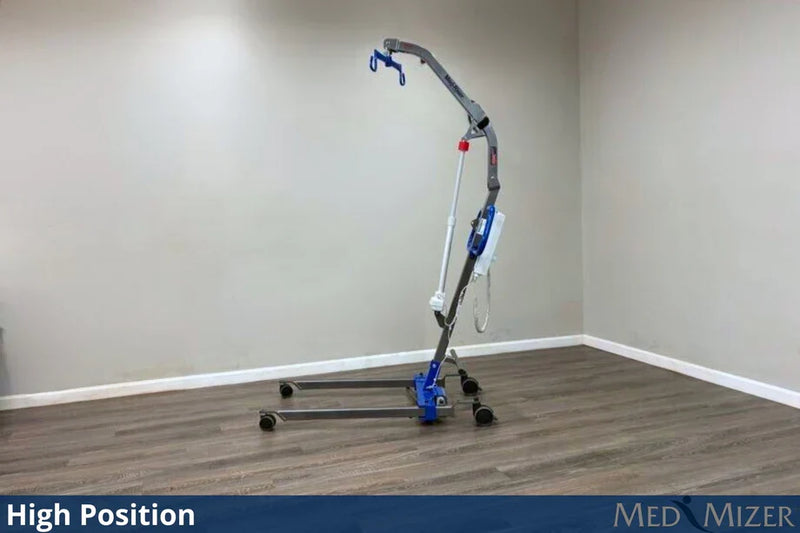 A high-position medical transfer chair with a wheeled base and curved lift arm, shown in a room with beige walls and wood-look flooring. The product has a gray frame with blue and white components.