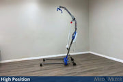 A high-position medical transfer chair with a wheeled base and curved lift arm, shown in a room with beige walls and wood-look flooring. The product has a gray frame with blue and white components.