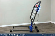 A medical transport lift in the folded position, shown on a wood-look floor against a beige wall. The product has a dark gray metal frame, blue hydraulic components, and black wheels.