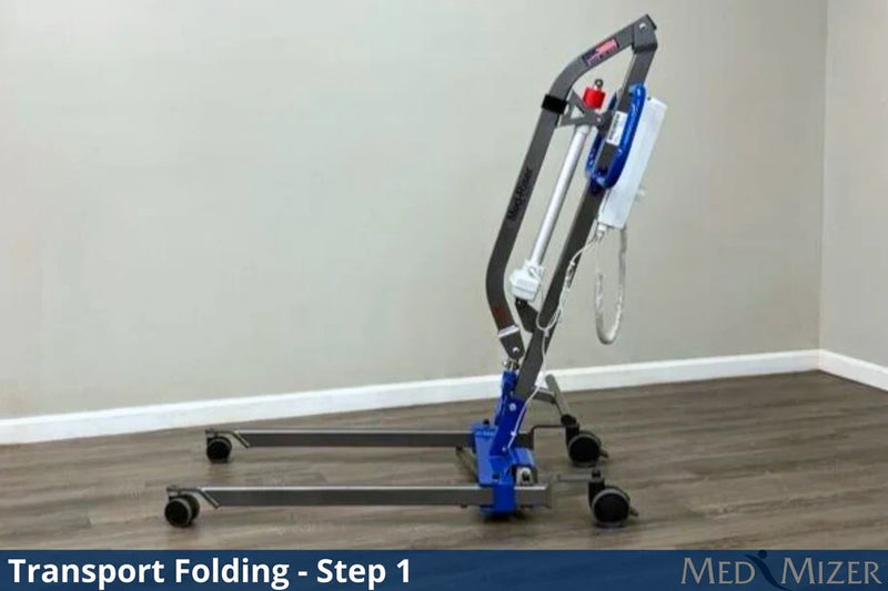 A medical transport lift in the folded position, shown on a wood-look floor against a beige wall. The product has a dark gray metal frame, blue hydraulic components, and black wheels.