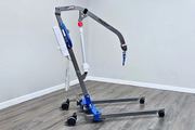 A medical lift chair with a gray metal frame, blue and white components, and black caster wheels, shown in a room with gray walls and wood-look flooring.