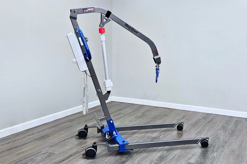 A medical lift chair with a gray metal frame, blue and white components, and black caster wheels, shown in a room with gray walls and wood-look flooring.