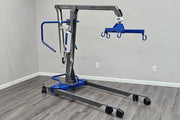 A blue and gray medical lift with a wheeled base and an overhead lifting arm, shown in a room with gray walls and wood-look flooring.