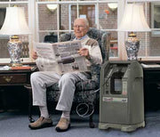 Man reading a newspaper in a chair next to a home oxygen concentrator.