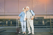 An elderly couple embracing and kissing in an airport terminal, with the woman pulling a rolling suitcase and the man holding a cane. The background features a large glass wall reflecting warm sunset light.