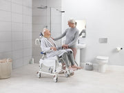 Woman using a shower chair in a bathroom with another person assisting her.