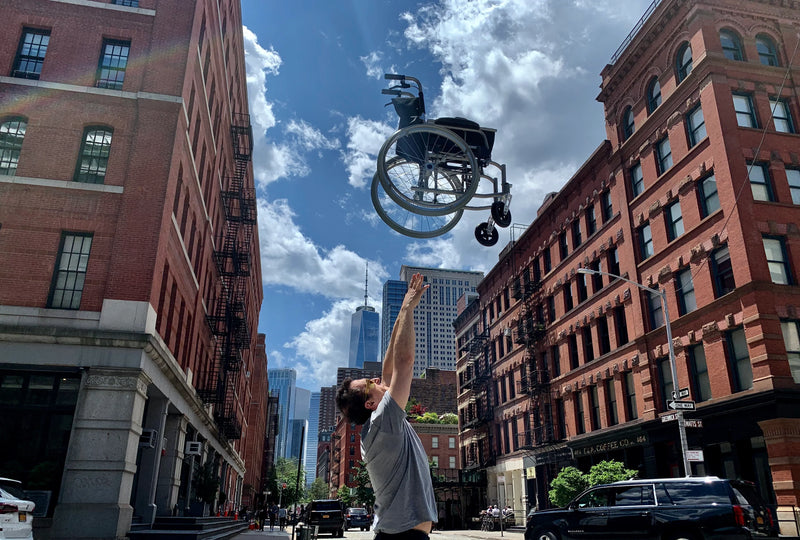 Person in a city street lifting a wheelchair upwards with buildings and blue sky in the background