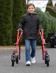 Child using a red walker on a paved path with greenery in the background