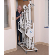 A white hospital bed lift on a wheeled base, shown being pushed by a man in a doorway inside a clinical room.
