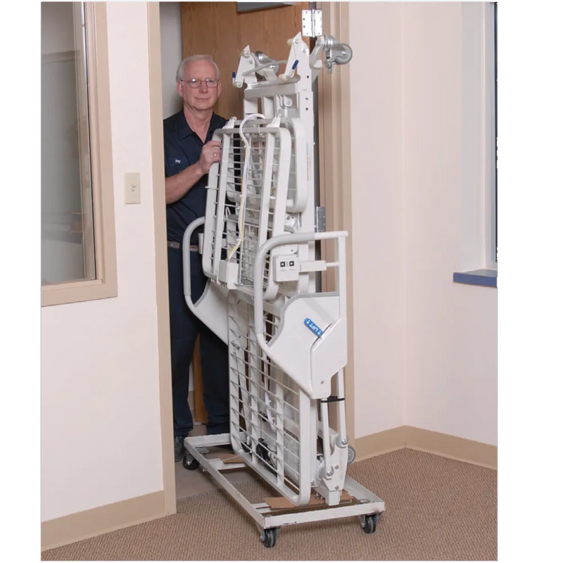 A white hospital bed lift on a wheeled base, shown being pushed by a man in a doorway inside a clinical room.