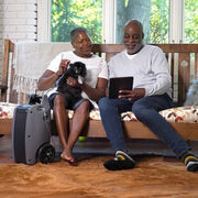 Two people sitting on a bench with a small dog, using a tablet, next to a portable oxygen concentrator.