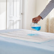 Person pouring blue liquid onto a mattress protector to demonstrate its water-resistant properties.